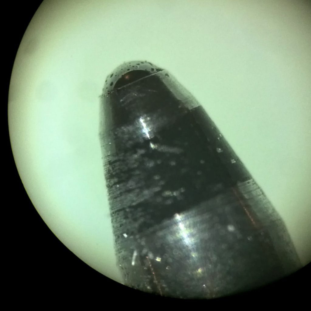 Spitze einer hochwertigen Kugelschreibermine unter dem Mikroskop. Die Kugel ist tatsächlich so präzise geformt, dass man darin die Spiegelung der Umgebung erkennen kann.